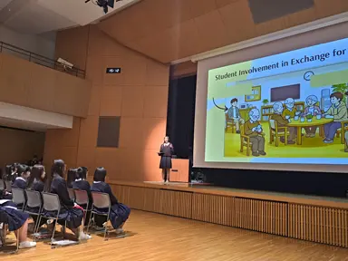 イラストの入ったプロジェクタ資料の前で女子生徒の発表を聞く生徒達