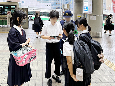 女子警察官に見守られ女子学生にチラシを配る男子生徒と女子生徒
