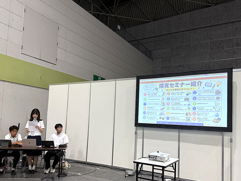 プロジェクタを映しながら本校高2女子生徒がマイクで発表